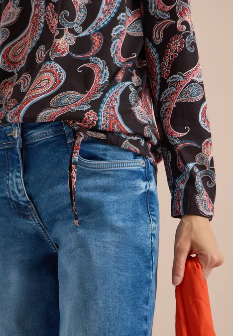 Ein Mensch trägt Paisley-Hemd, blaue Jeans und hält ein oranges Tuch in der rechten Hand.
