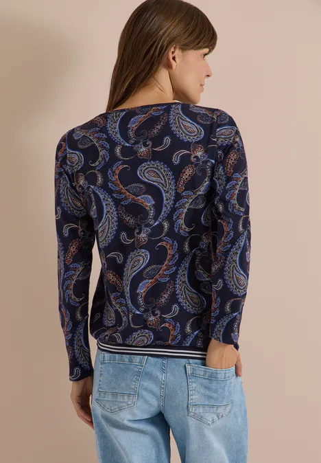 Rückenansicht einer Frau in dunkelblauem, paisley-bedrucktem Langarmshirt und hellblauen Jeans.