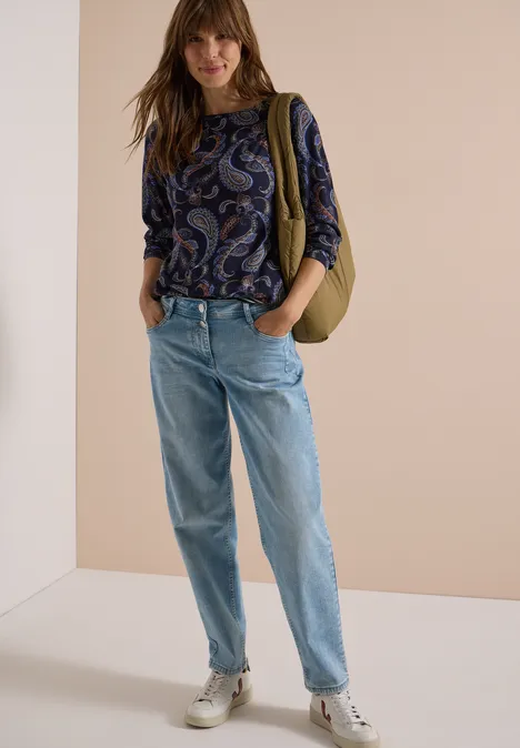 Frau in Paisley-Bluse, weiten Jeans, weißen Sneakers und grüner Umhängetasche vor neutralem Hintergrund.