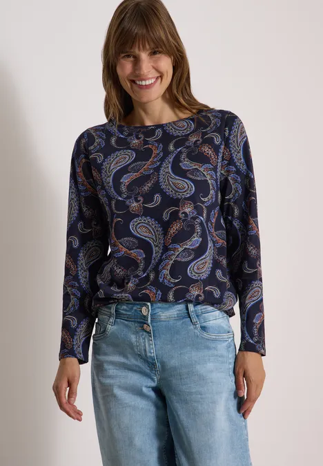 Lächelnde Frau mit blauem Paisley-Oberteil und hellblauen Jeans vor neutralem Hintergrund