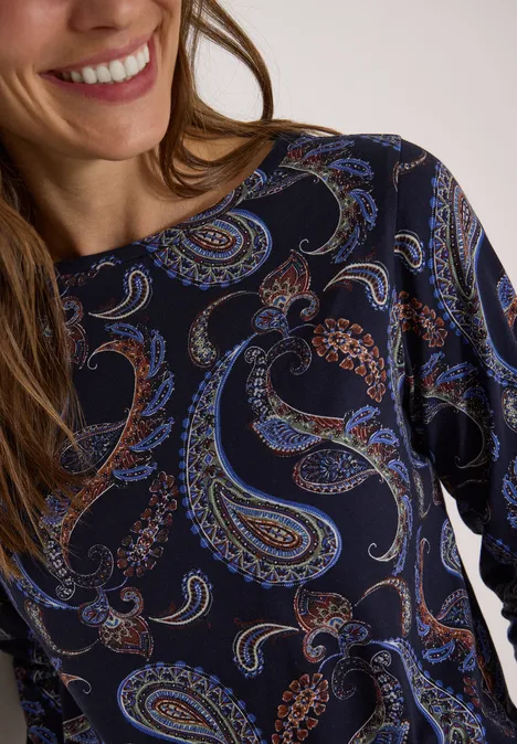 Nahaufnahme einer Frau im dunkelblauen Shirt mit buntem Paisley-Muster und leichtem Lächeln.