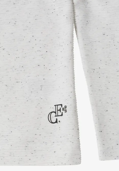 Nahaufnahme eines cremeweißen, melierten Pullovers mit dezentem schwarzen Logo am Saum.