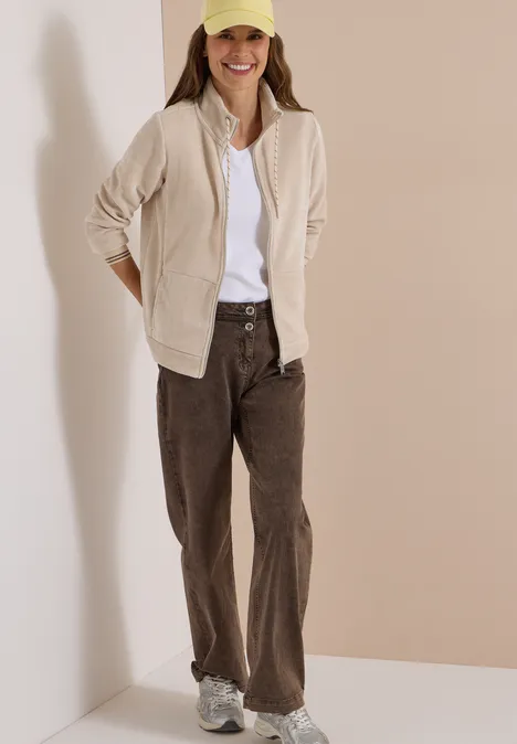 Junge Frau in heller Jacke, weißem Shirt, braunen Cordhosen, Sneakern und gelber Mütze vor beigefarbenem Hintergrund.