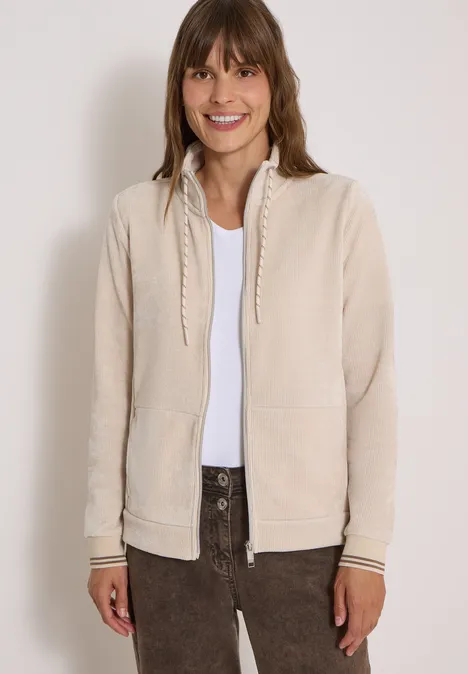 Eine lächelnde Frau in beiger Strickjacke, weißem Shirt und dunklen Hosen vor hellem Hintergrund.