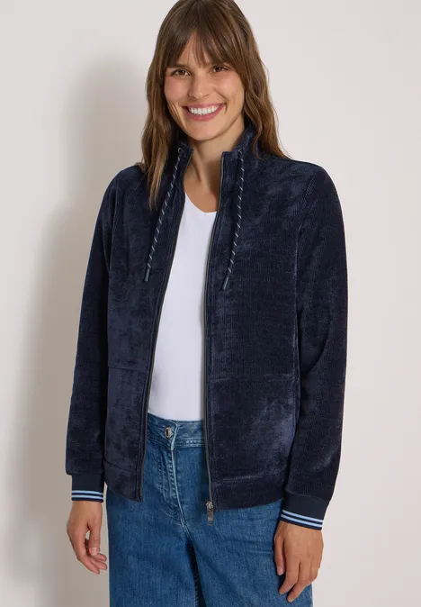 Frau mit mittellangem braunem Haar trägt eine dunkelblaue Plüsch-Jacke, weiße Bluse und Jeans, lächelnd.
