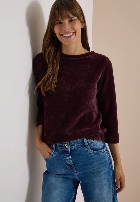T-shirt à manches 3/4 en velours côtelé avec encolure bateau berry red Jeune femme souriante appuyée contre un mur, portant un pull violet et un jean bleu.