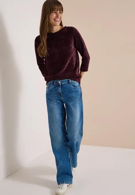 T-shirt à manches 3/4 en velours côtelé avec encolure bateau berry red Jeune femme souriante en jean large bleu et pull bordeaux, marchant dans un espace épuré et lumineux.