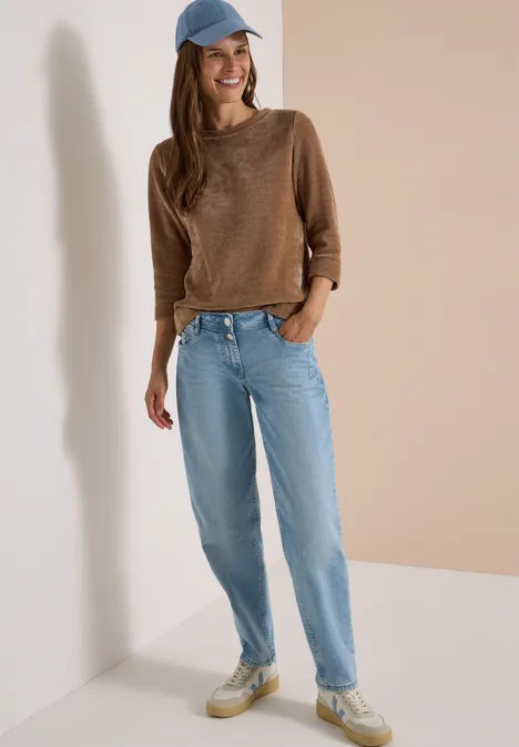 T-shirt à manches 3/4 en velours côtelé avec encolure bateau creamy mocha Femme appuyée contre un mur, pull marron, jean bleu clair, baskets blanches et casquette bleue.