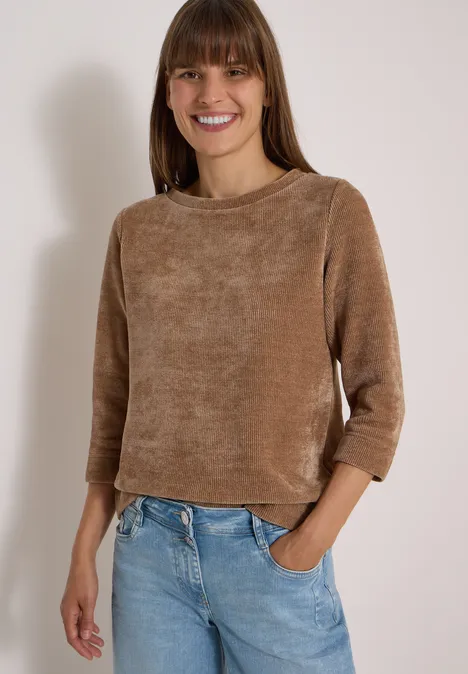 T-shirt à manches 3/4 en velours côtelé avec encolure bateau creamy mocha Une femme souriante porte un pull chenille marron et un jean bleu clair, mains en poche, sur fond clair.