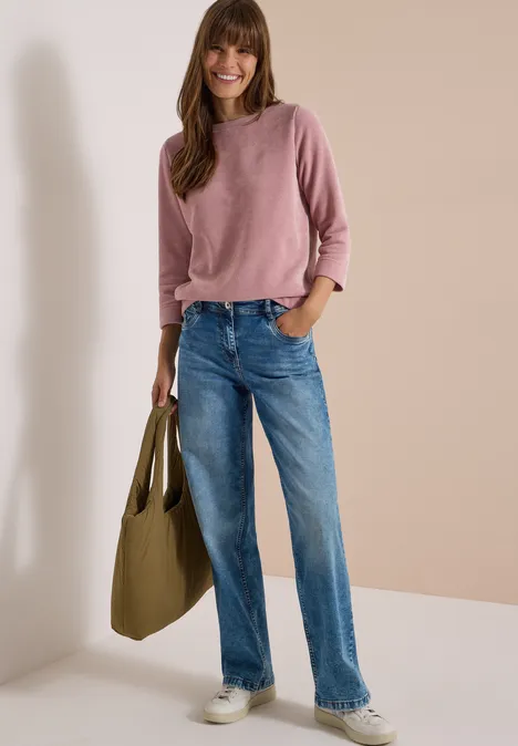Eine Frau in rosa Pullover, blauen geraden Jeans und weißen Sneakern, trägt eine grüne Tasche.