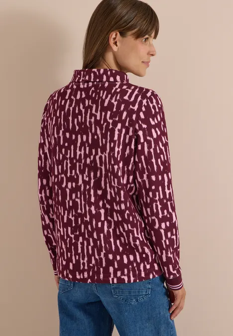 Frau mit braunen Haaren, in lila-pink gemustertem Langarmshirt und blauen Jeans, seitlich stehend