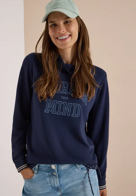 Junge Frau in Jeans, dunkelblauem Sweatshirt mit 'OPEN MIND'-Aufdruck und hellgrüner Cap lächelt vor hellem Hintergrund.