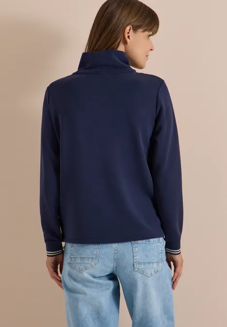 Frau von hinten in dunkelblauer Polo-Strickjacke mit Stehkragen und heller Jeans vor beigem Hintergrund.