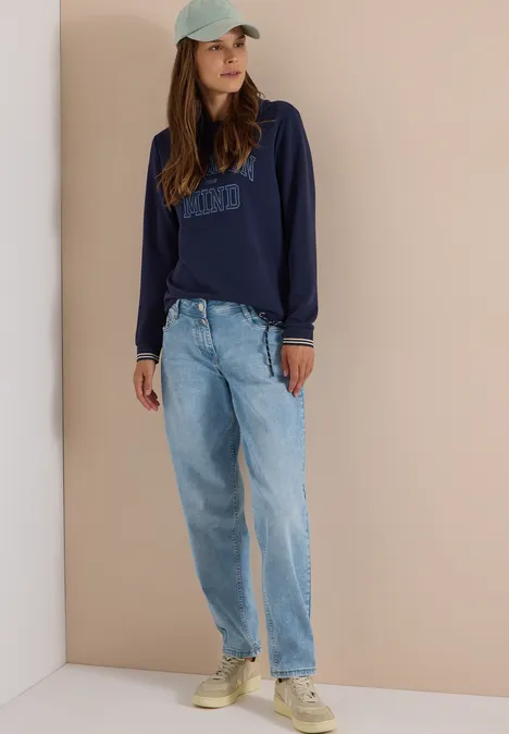 Frau mit dunkelblauem Sweatshirt, lockeren hellblauen Jeans, hellen Sneakern und mintgrüner Cap vor beigem Hintergrund.