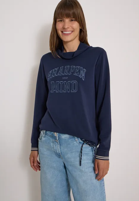 Lächelnde Frau in marineblauem Sweatshirt mit Aufdruck und hellen Jeans.