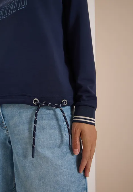 Halber Torso: dunkelblaues Sweatshirt mit Kordelzug, weiß gestreiften Bündchen, hellblaue Jeans, eine Hand.