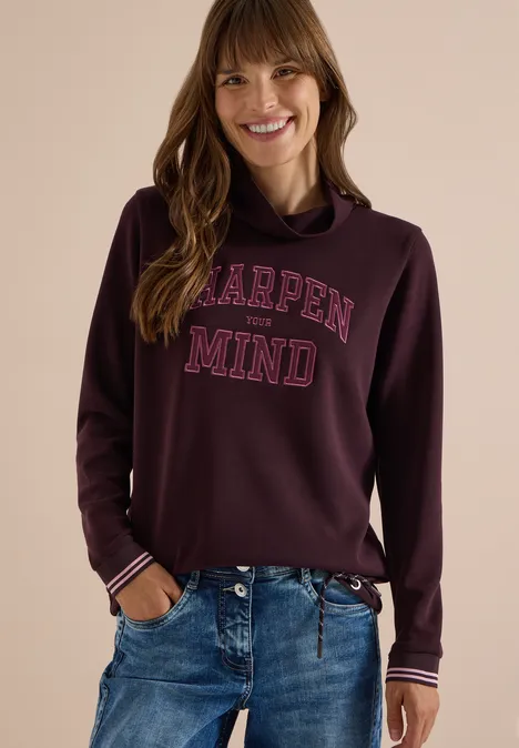 Frau in burgunderrotem Kapuzenpullover mit 'Sharpen Mind'-Aufschrift, lächelnd in Jeans.