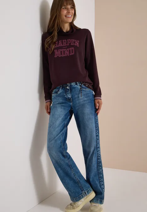 Frau lehnt an Wand, lässiges bordeauxfarbenes Sweatshirt mit Schriftzug und weite blaue Jeans.