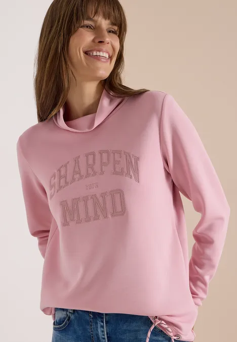 Eine Frau trägt ein hellrosa Sweatshirt mit hohem Kragen und Schriftzug.
