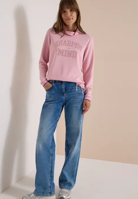 Eine Frau in rosa Kapuzenpullover und blauen Jeans vor neutralem Hintergrund.