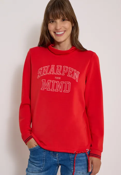 Frau im roten Sweatshirt mit Aufdruck 'Sharpen Your Mind' und blauer Jeans lächelt in die Kamera.