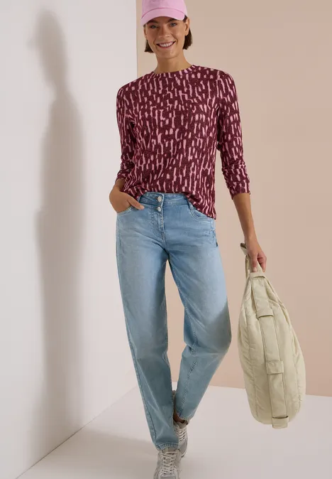 Frau mit lila gemustertem Shirt, hellen Jeans, Cap und gestepptem Beutel, lächelnd an einer Wand entlang.