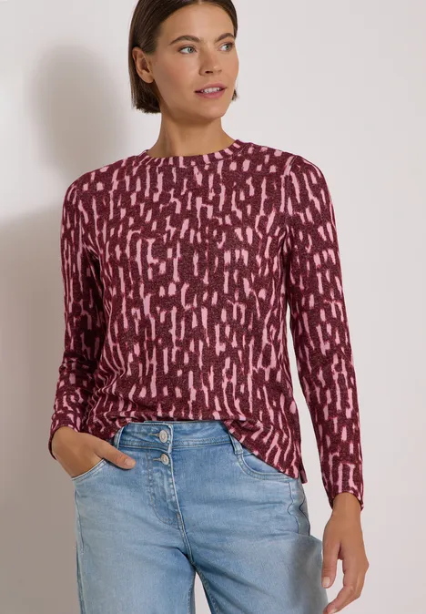 Frau trägt bordeauxfarbenes, gemustertes Langarmshirt und helle Jeans vor neutralem Hintergrund.