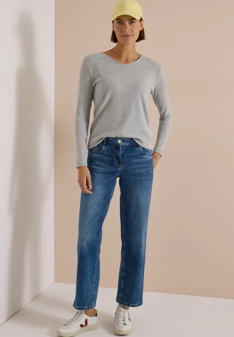 Frau in blauer Jeans, grauem Langarmshirt, weißen Sneakers und gelber Cap vor neutralem Hintergrund.