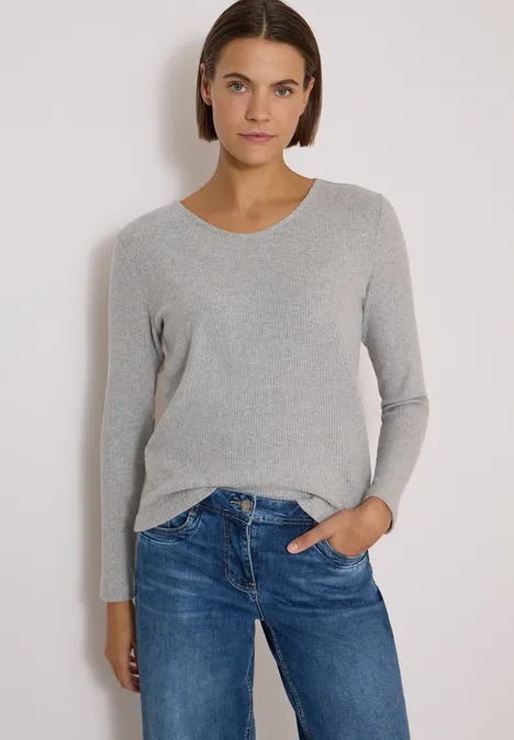 Frau in grauem Pullover und blauen Jeans vor hellem Hintergrund, blickt direkt in die Kamera.