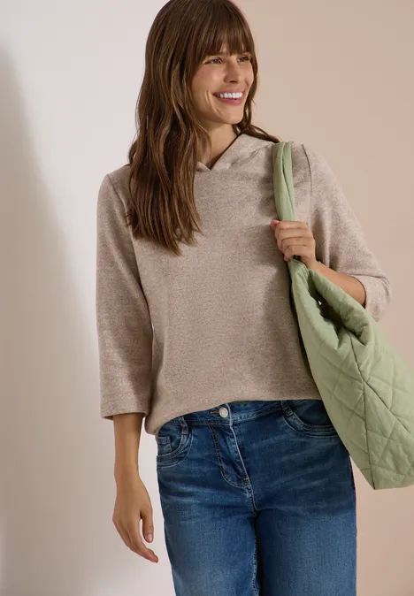 Femme souriante en pull à capuche beige, jean bleu et sac vert.
