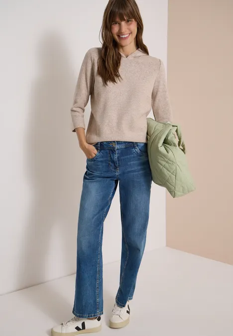Une femme souriante porte un pull beige, un jean bleu, des baskets blanches et tient une veste vert clair.
