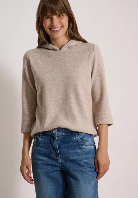 Une femme souriante porte un pull à capuche beige clair et un jean bleu.