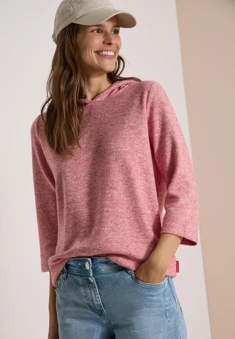 Femme souriante en sweat à capuche rose, manches 3/4, jean et casquette.
