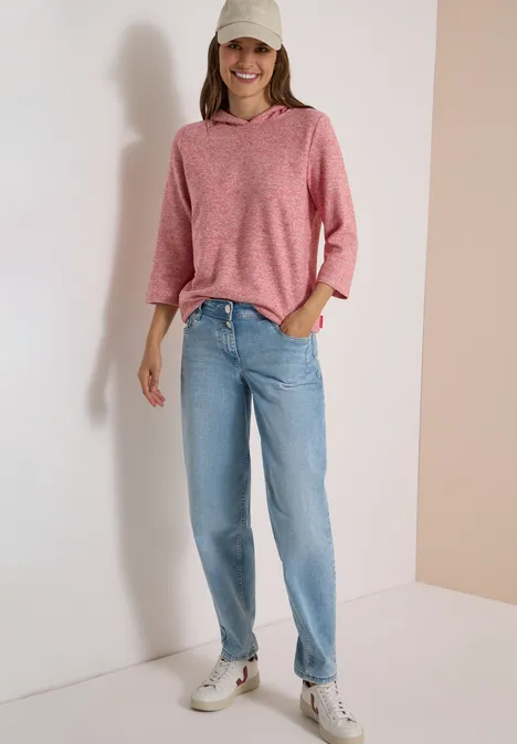Femme en hoodie rose, jean bleu et baskets blanches devant un fond neutre.