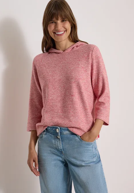 Femme souriante vêtue d'un pull à capuche rose et d'un jean bleu, posant détendue les mains dans les poches.