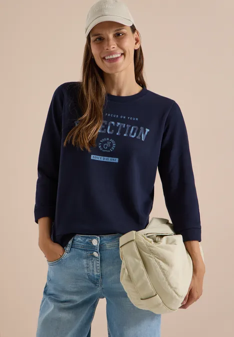 T-shirt à manches 3/4, col rond et inscription urban dark blue Une femme souriante porte un sweat-shirt bleu marine avec inscription, un jean et un grand sac à bandoulière.
