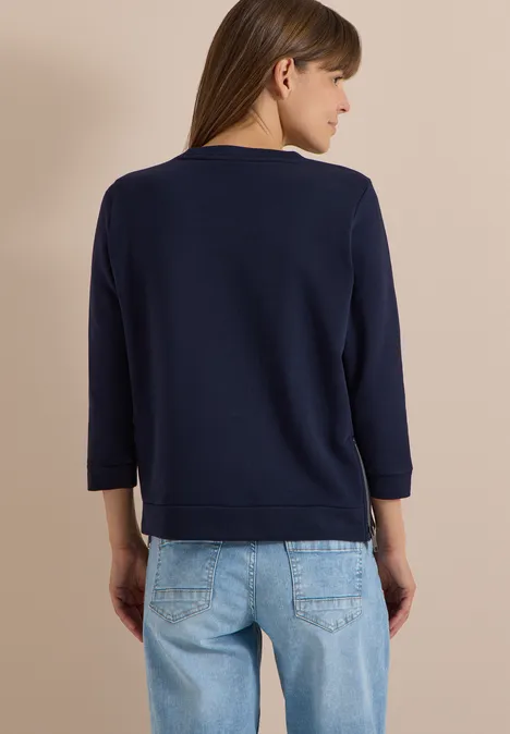 T-shirt à manches 3/4, col rond et inscription urban dark blue Vue de dos d’une femme en haut bleu marine à manches longues et jean bleu clair sur fond beige.
