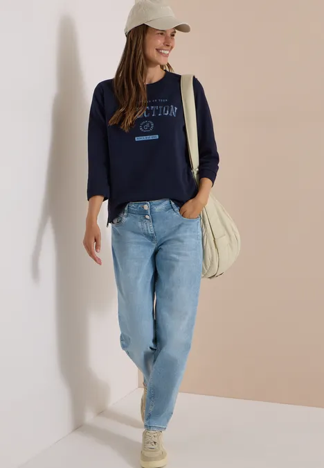 T-shirt à manches 3/4, col rond et inscription urban dark blue Une jeune femme en casquette, sweat bleu marine, jean clair, baskets et sac devant un mur bicolore
