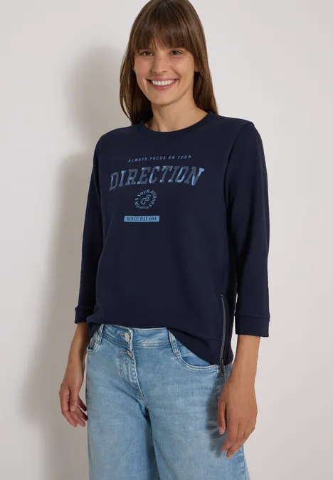 T-shirt à manches 3/4, col rond et inscription urban dark blue Femme souriante en jean et t-shirt bleu marine 'Direction', posant sur fond clair.