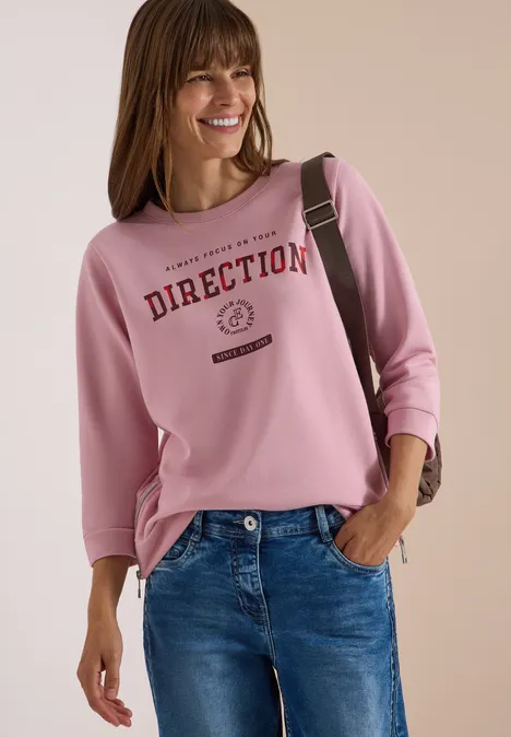 T-shirt à manches 3/4, col rond et inscription mellow rose Femme souriante en sweat rose avec inscription 'DIRECTION', jean bleu et sac à l’épaule.