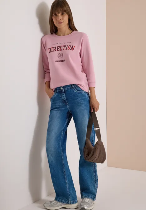 T-shirt à manches 3/4, col rond et inscription mellow rose Femme en sweat rose imprimé, jeans bleus et sac marron porté à l'épaule devant un fond neutre.