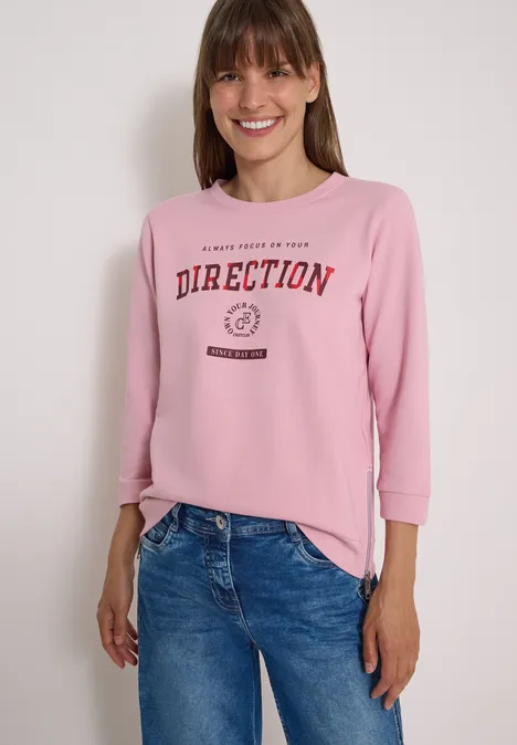 T-shirt à manches 3/4, col rond et inscription mellow rose Femme souriante portant un sweat rose avec l’inscription « DIRECTION » et un jean bleu.