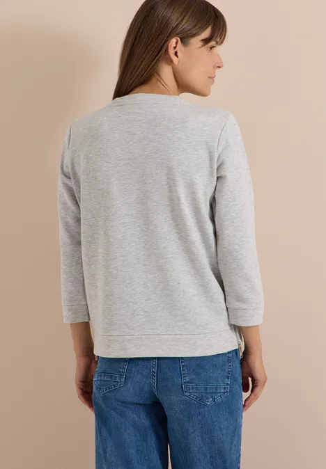 Frau von hinten in grauem Sweatshirt mit 3/4-Ärmeln und blauen Jeans vor hellem Hintergrund.