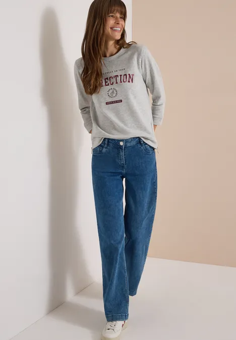 Eine lachende Frau in grauem Sweatshirt und blauen, gerade geschnittenen Jeans vor hellem Hintergrund.