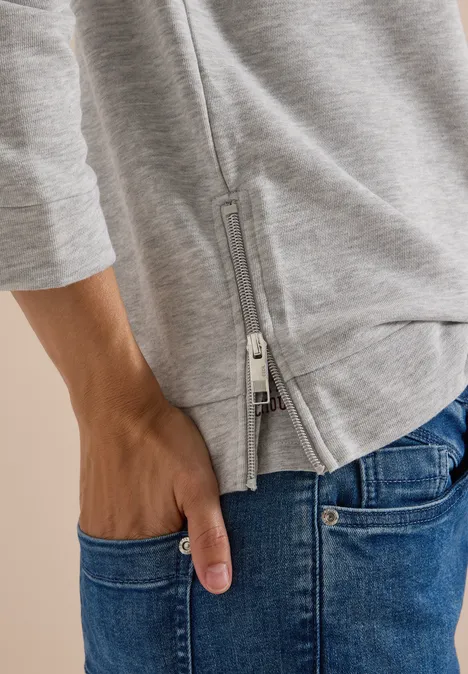 Hand in Jeans-Tasche, darüber graues Oberteil mit seitlichem Reißverschluss.