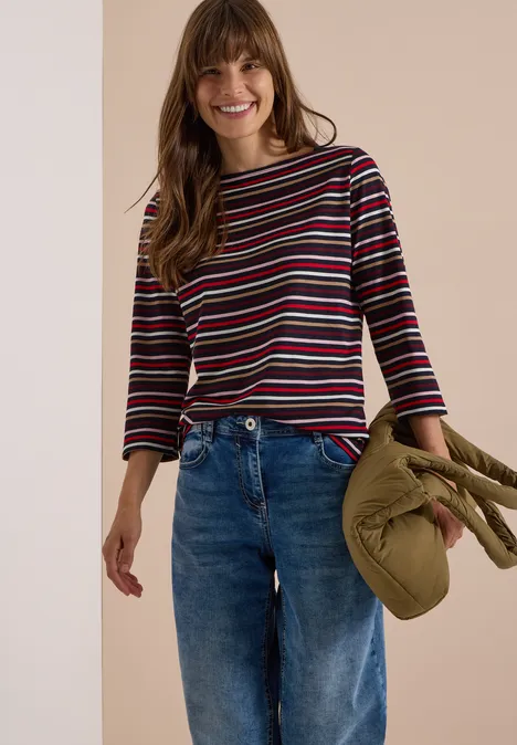 Frau in gestreiftem Langarmshirt und Jeans, hält grünen Mantel in der Hand, lächelt entspannt.