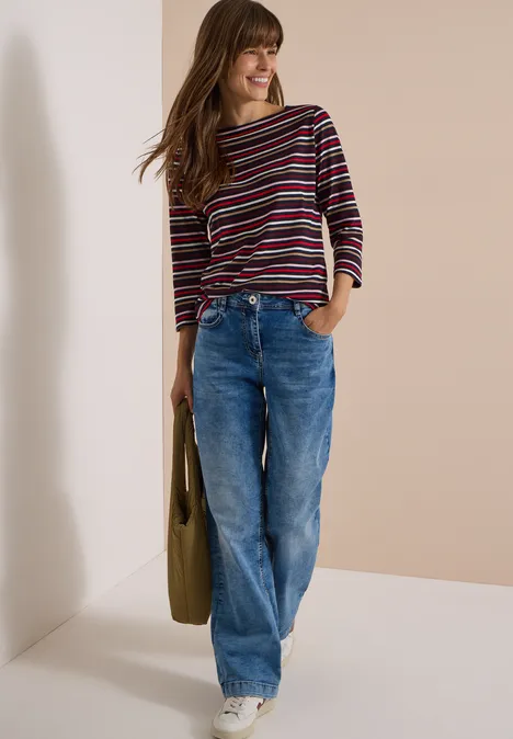 Junge Frau in gestreiftem Pullover, weiten Jeans und weißen Sneakers lehnt lächelnd an einer Wand, trägt Tasche.
