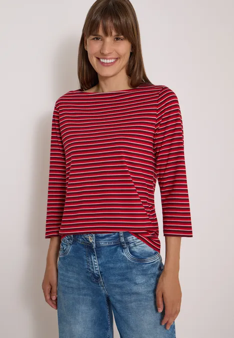 Frau mit rot-dunkelblau gestreiftem Langarmshirt und Jeans lächelnd vor hellem Hintergrund.