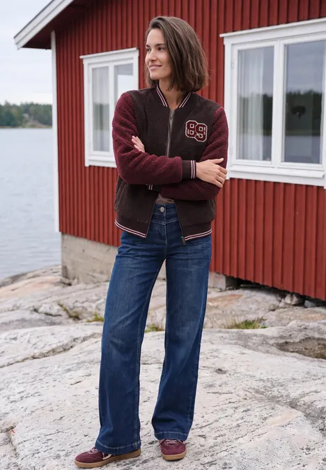 Frau in Jeans und Collegejacke steht vor einem roten Haus am See auf Felsen.