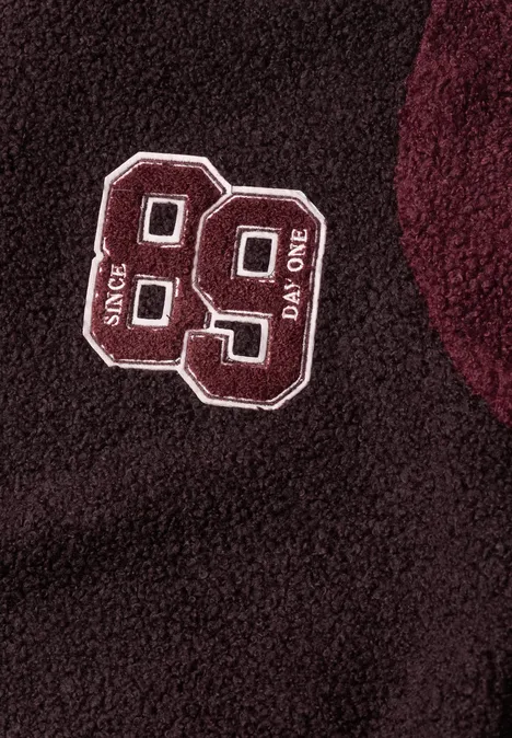Dunkelrotes Fleece-Kleidungsstück mit gesticktem weißem Patch '99' und Schriftzügen 'SINCE' und 'DAY ONE'.